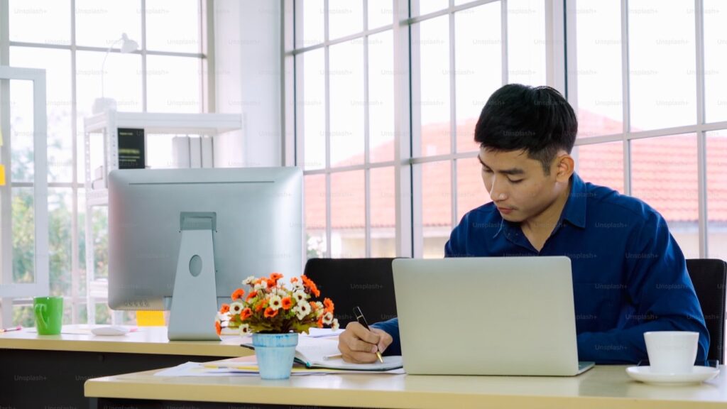 FXの基礎知識を身につけるため、ノートパソコンの前で真剣にメモを取り勉強する大学生の男性。